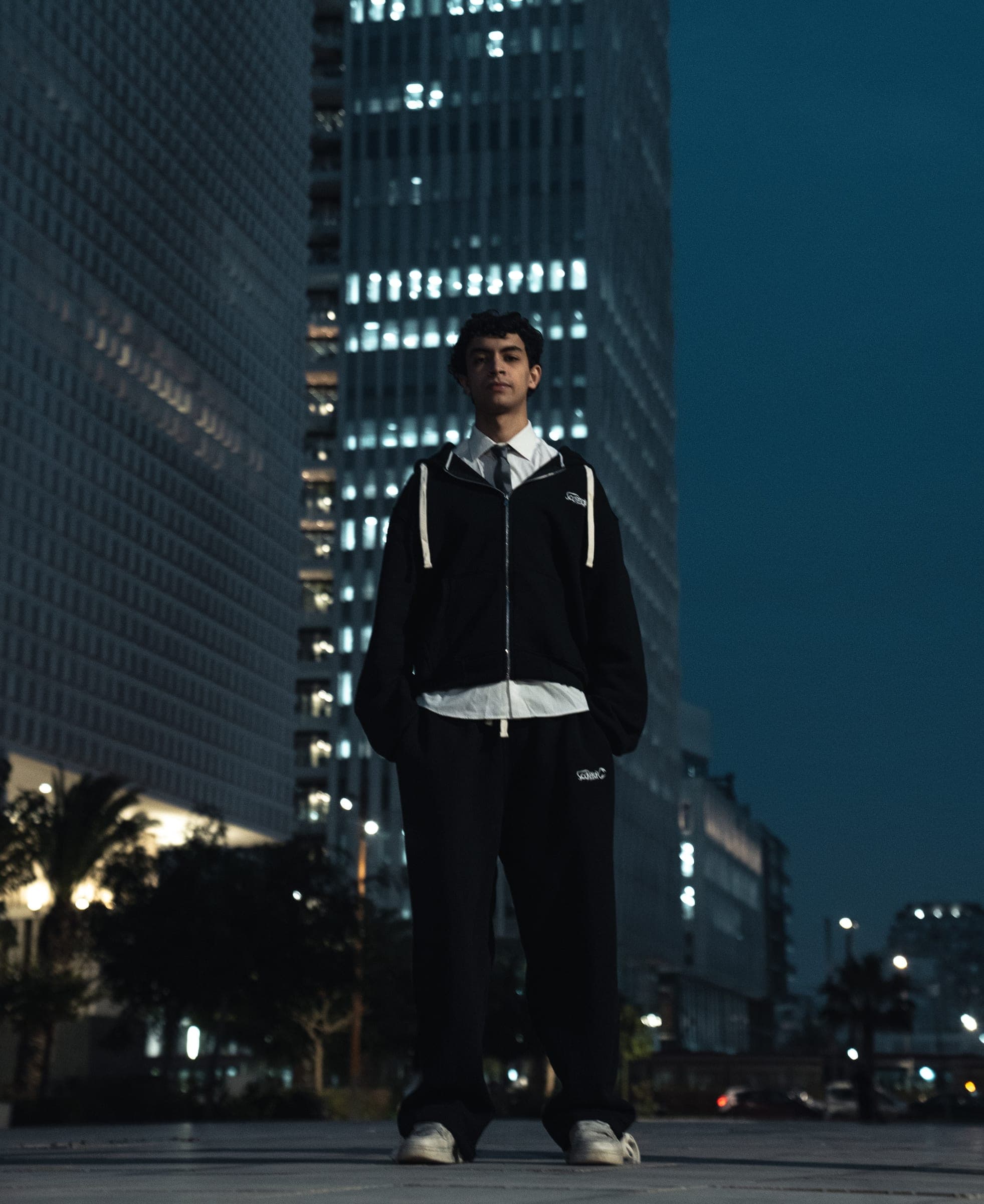 A crew member at night in the black zip-up hoodie and wide pants, lit office towers behind