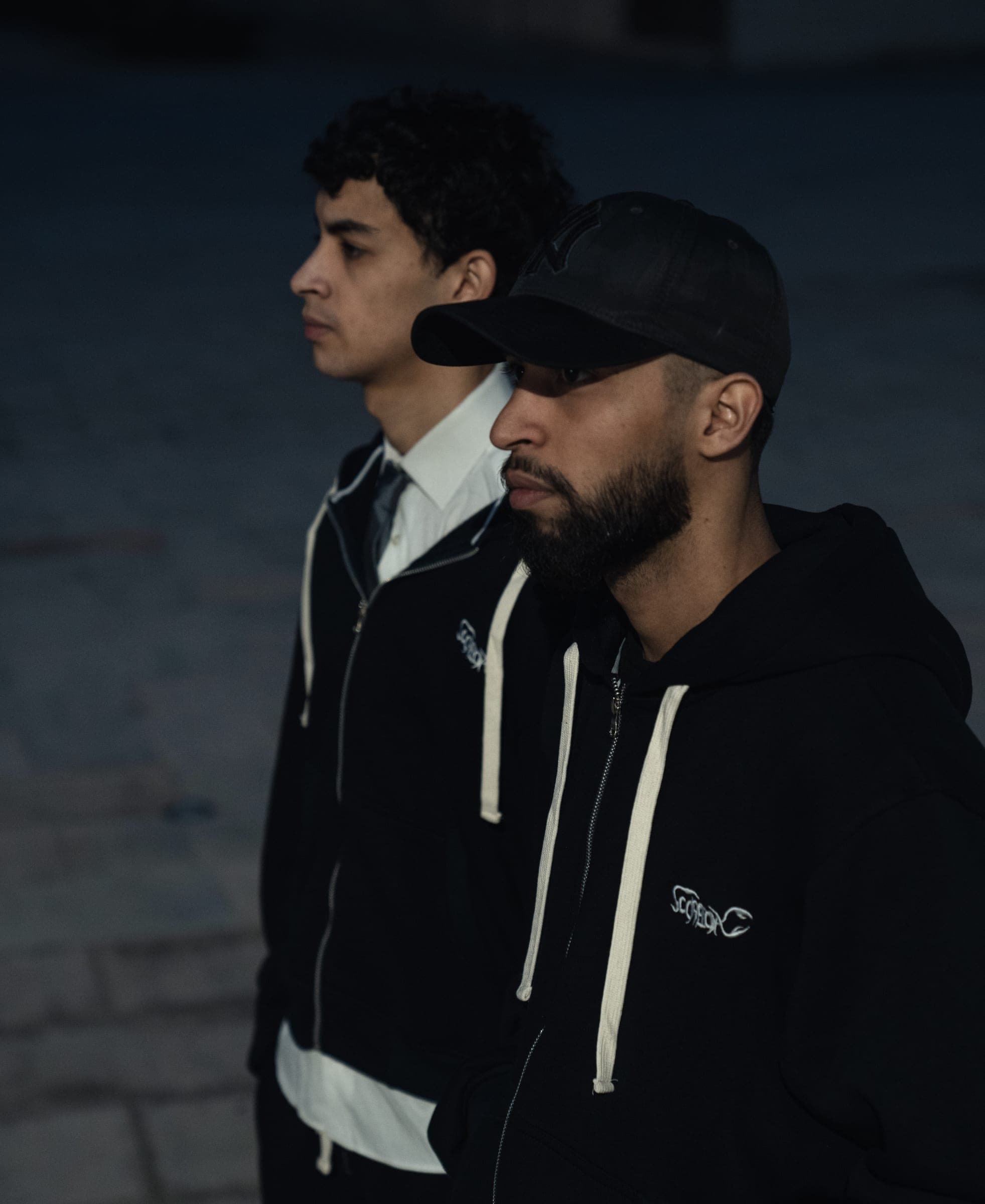 Two crew members at night in zip-up hoodies over white shirts, one in a black cap