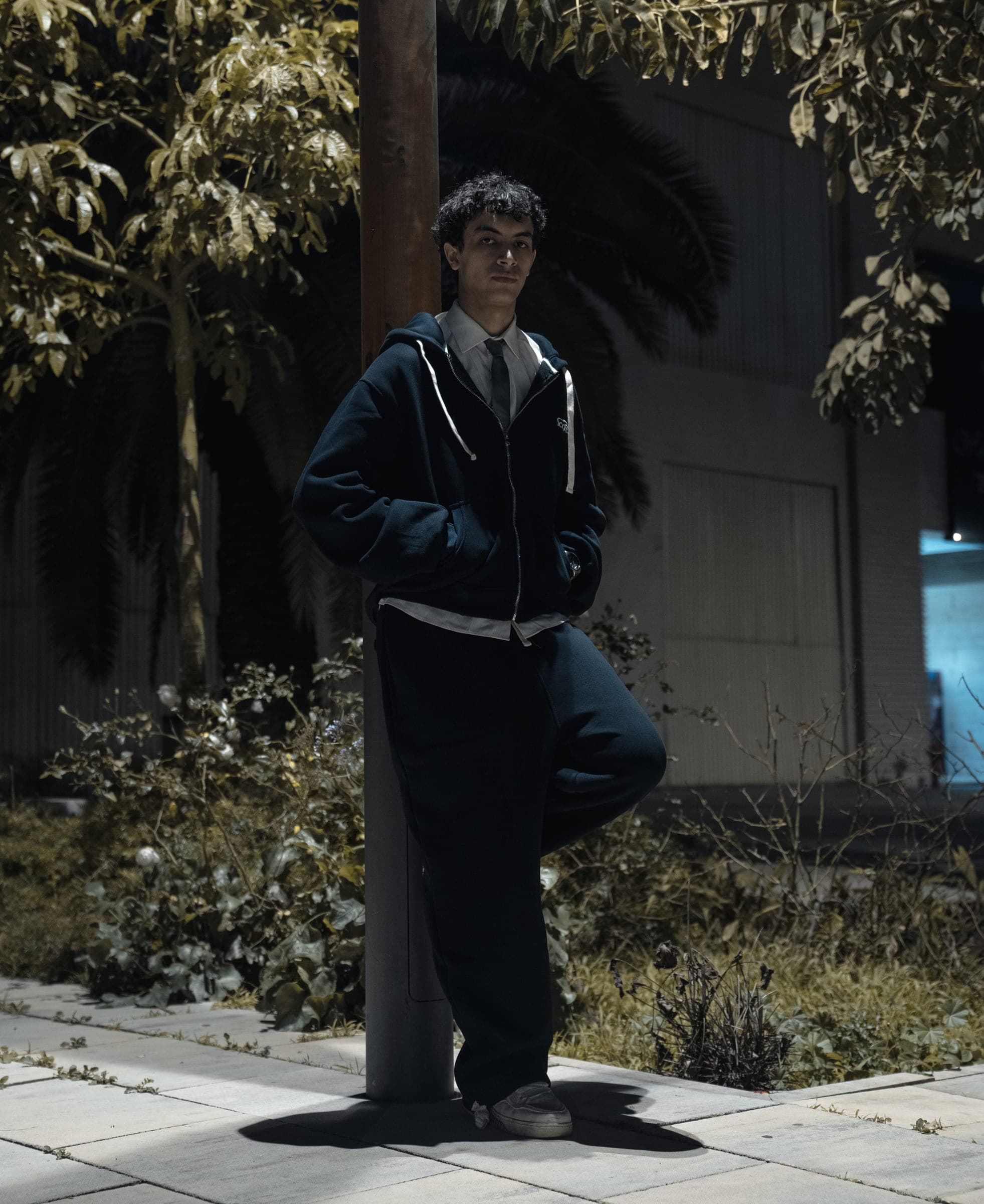 A crew member leaning on a post between palms in the zip-up hoodie and pants